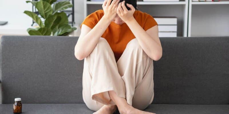 Naine istub diivanil ja hoiab kätega peast kinni, kõrval ravimipurk